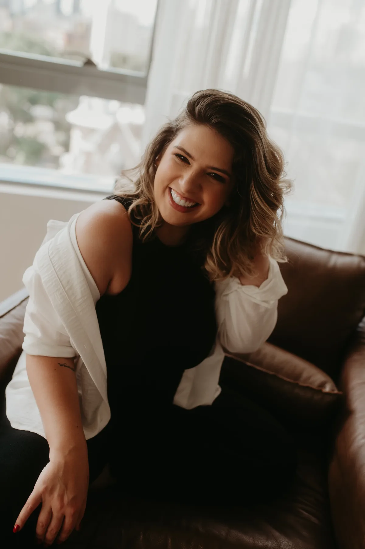 Aline Candatten sentada numa poltrona de couro, sorrindo para a câmera, com a luz da janela atrás